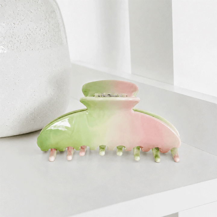A glossy hair claw clip with a pastel green and pink gradient, resting on a white shelf next to a textured white vase.