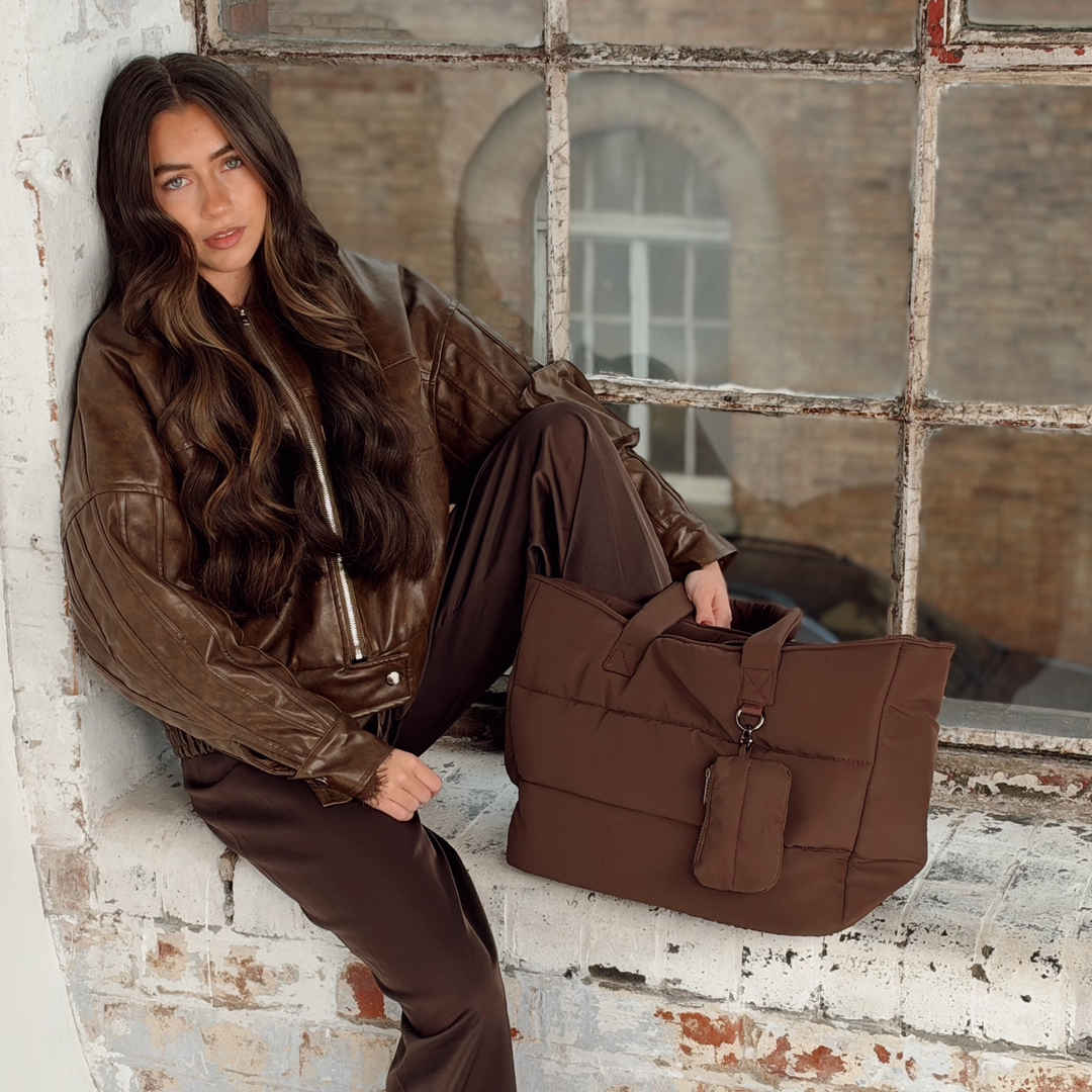 Woman with long brunette hair sat on a industrial brick window sill setting. Next to her is the Coconut Lane Main Carry choc brown puffer bag. Attached on the external d-ring is the matching mini keyring bag. 