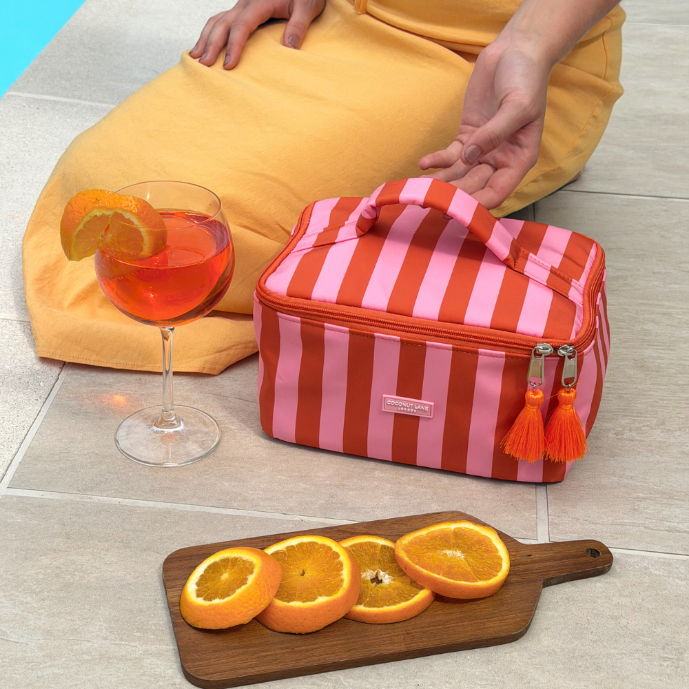 Orange and pink striped cooler bag with tassels, a glass of aperol spritz, and a cutting board with oranges on a tiled floor.