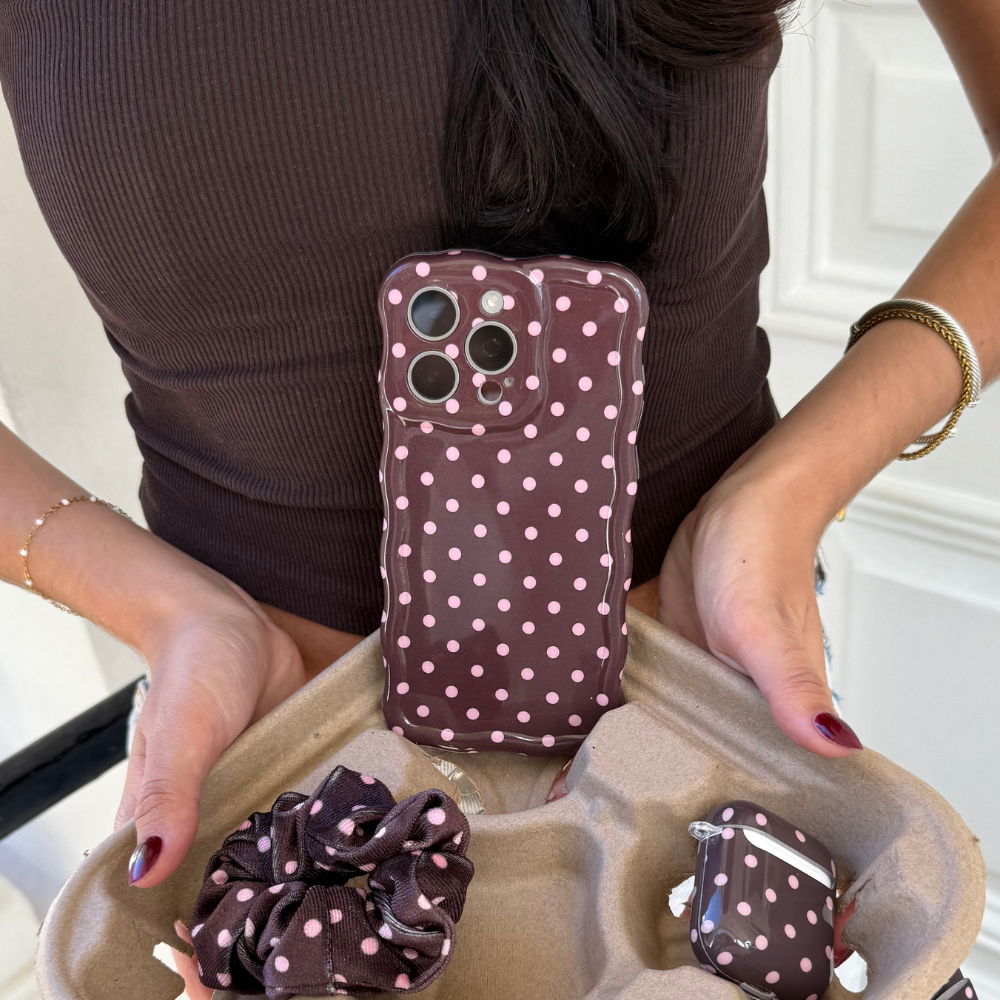 Woman holding wavy phone case, brown polka dot scrunchie, and matching AirPods case from Coconut Lane.