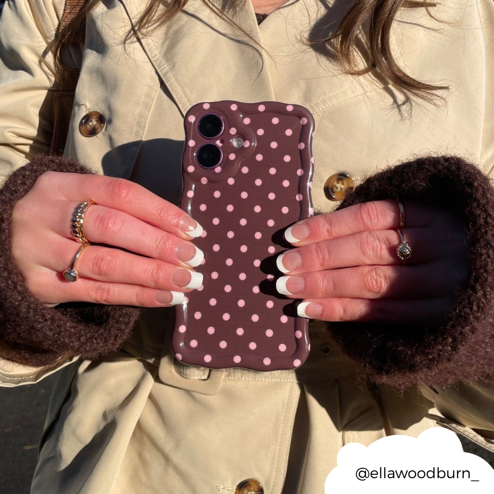 Hand with French tip nails holds a brown polka dot wavy phone case, paired with a beige coat.