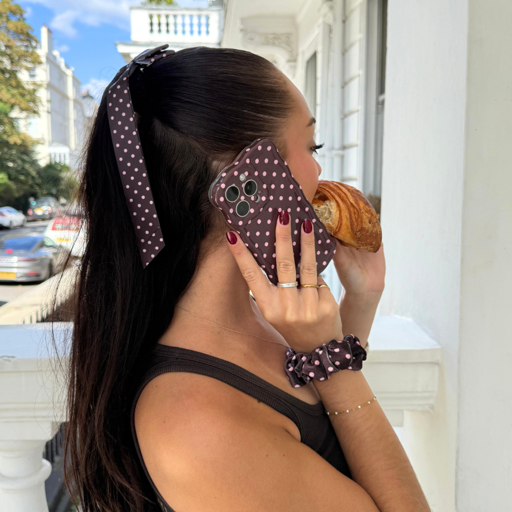 Woman holding a brown polka dot wavy phone case eats a croissant on her balcony.