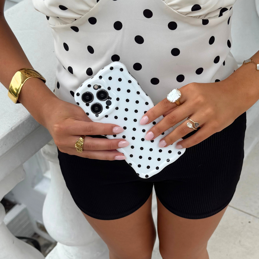 Woman in a polka dot top holding a white Coconut Lane phone case with black wavy polka dots, wearing black shorts.