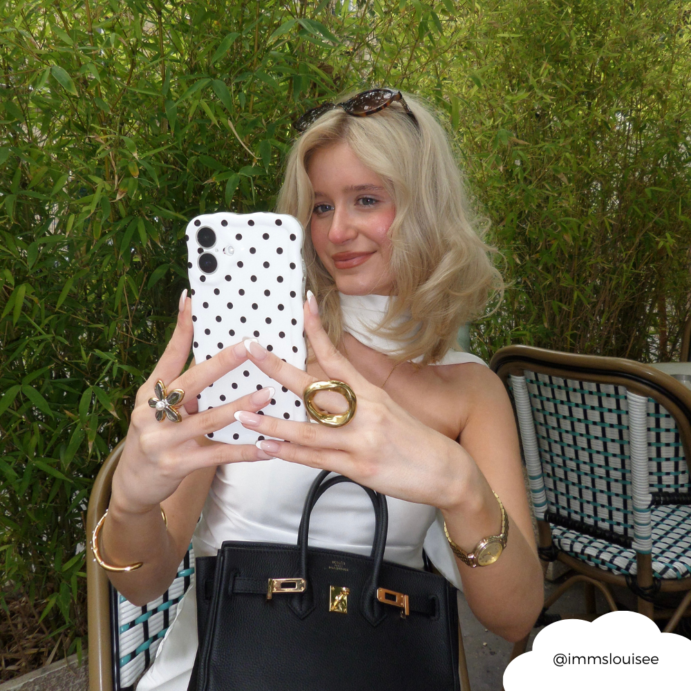 Blonde woman at an outdoor café holding a white polka dot wavy phone case and a black bag.
