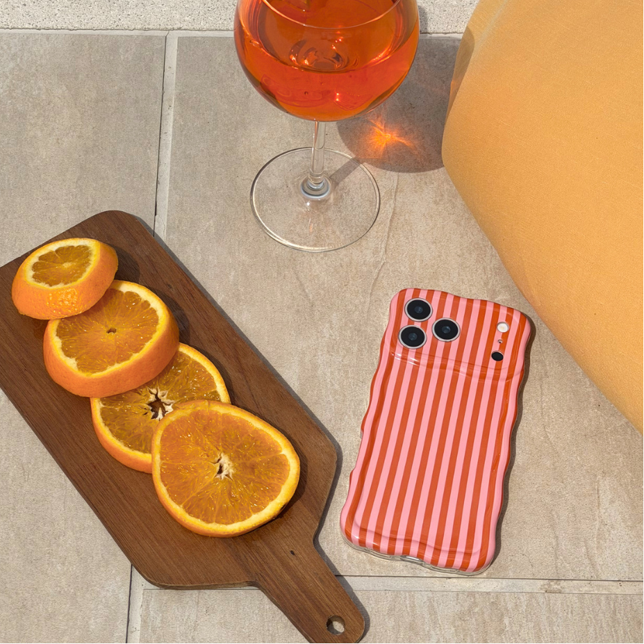 Phone case with orange and pink striped pattern next to sliced oranges and a glass of orange juice on a stone surface.