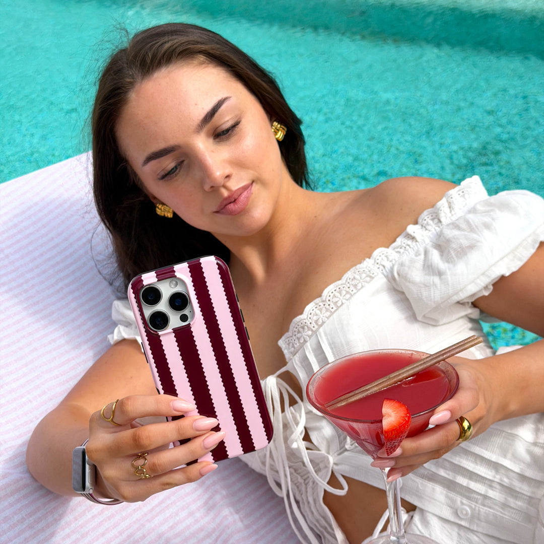 Girl holding a blushberry striped phone case, with strawberry daiquiri by the pool 