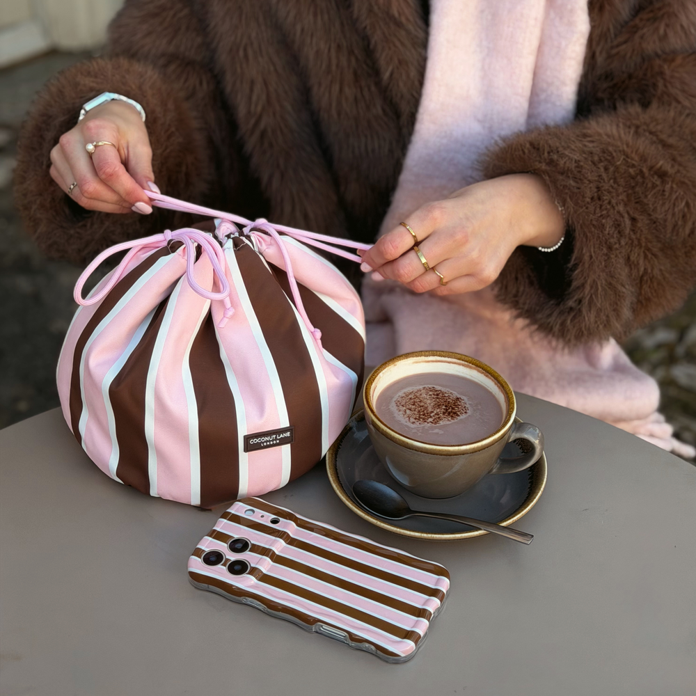 Woman wearing a baby pink scarf and brown faux fur jacket, holding a drawstring toiletry bag featuring chocolate brown, baby pink and white stripes with pink drawstring closure. Sat on the table next to this is the matching wavy phone case.