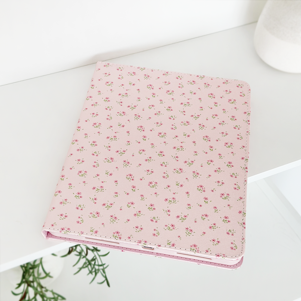 A pink notebook adorned with a delicate floral pattern rests on a white surface. A plant with lush green leaves is partially visible in the corner, and to the right, a white cylindrical object, likely a vase, is seen in the background. The setting perfectly complements my Coconut Lane Ditsy Floral Pink iPad Case.