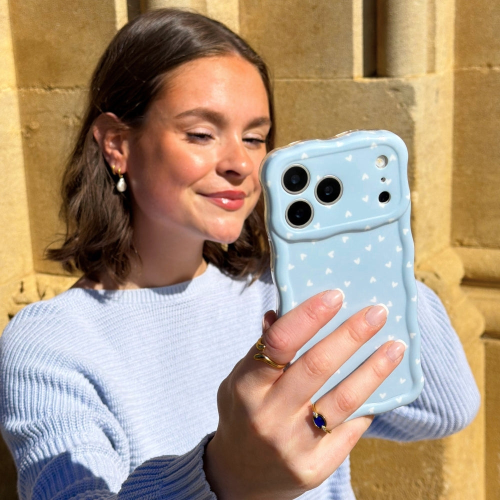 Woman holding powder blue hearts phone case against a stone wall in the sunshine with blue jumper