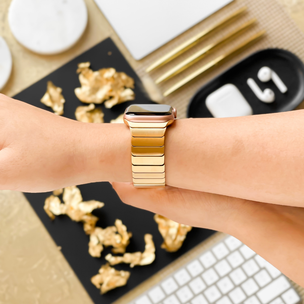 A person showcasing a Coconut Lane Slinky Apple Watch Strap in gold on their wrist. The backdrop includes an assortment of golden accessories, a pair of white AirPods in a black case, a white keyboard, and other meticulously arranged small white and gold items.