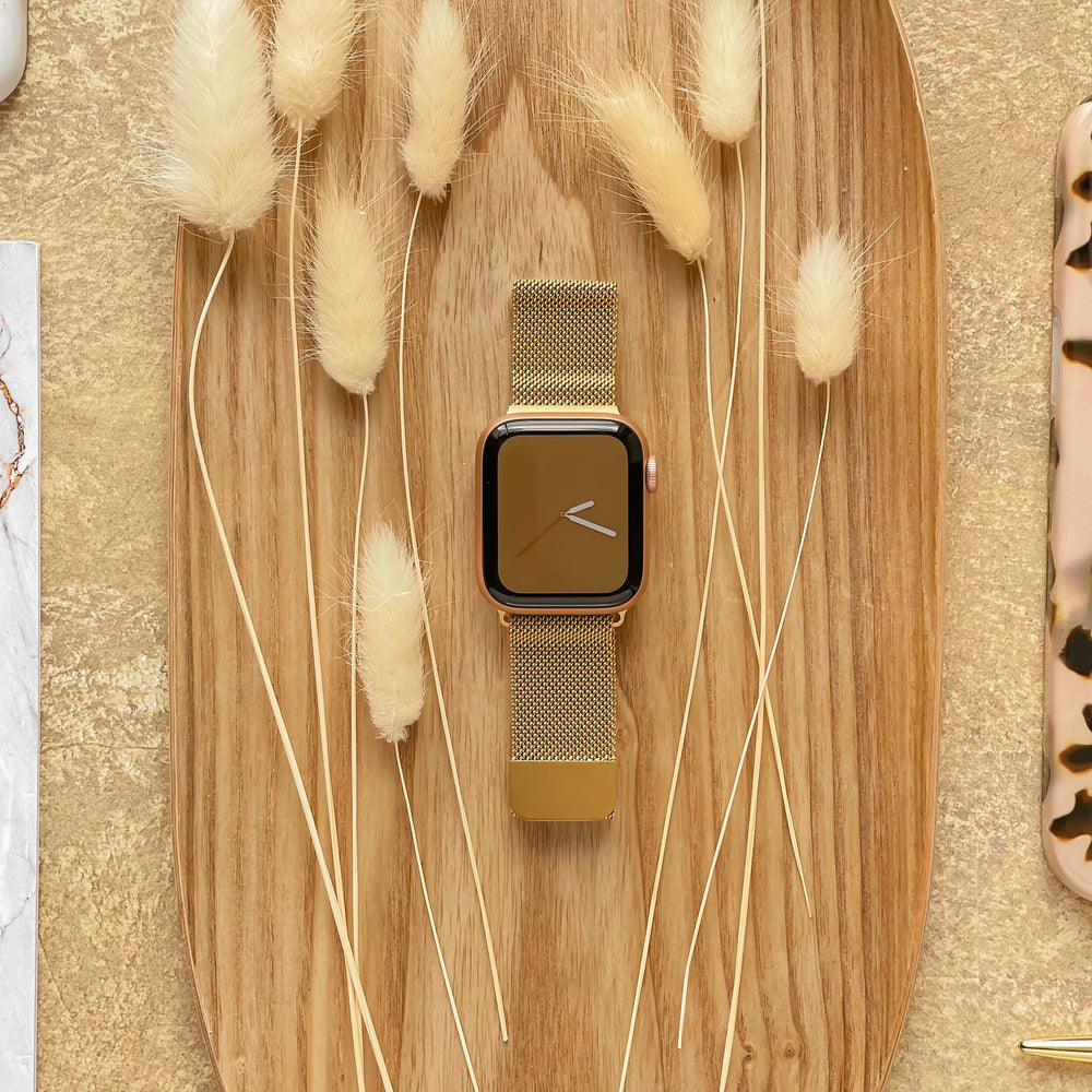 A gold Coconut Lane Magnetic Chain Apple Watch Strap is placed on an oval wooden tray, surrounded by dried beige fluffy grasses. The background features a soft golden-textured surface and parts of other items, including a marble-patterned object and a tortoiseshell object.